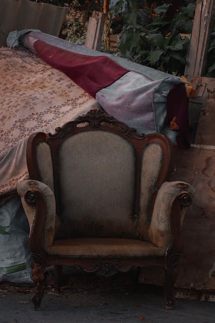 A vintage armchair with a carved wooden frame and upholstered back and seat, positioned in a room with wooden flooring. Behind the armchair, a large rolled-up carpet with a burgundy and grey geometric pattern rests on a wooden surface. To the left, part of a patterned fabric-covered table or surface is visible, and in the background, there are green leafy plants and a wooden wall or partition. The scene appears to be part of a home relocation or packing process, with furniture and items prepared for moving, consistent with house removals services offered by Man and Van Paddington, as seen on the webpage about bulky waste removal in Paddington W2.
