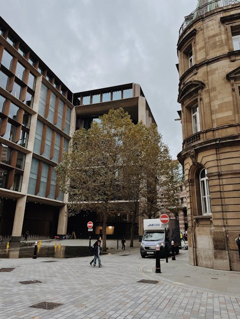 Exterior view of a city street in Paddington during daytime, featuring a mix of modern and historic architecture. On the left, a contemporary building with large glass windows and a concrete facade is visible, while on the right, an ornate, curved stone building with detailed masonry and arched windows adds historic character. A large tree with sparse foliage occupies the center, partially obscuring the buildings behind it. At street level, a grey moving van with the logo of Man and Van Paddington is parked close to the curb, with the rear doors open, indicating a loading or unloading process. Several cardboard boxes and wrapped furniture items can be seen inside the van and on the pavement nearby, suggesting furniture transport and home relocation activities. A few pedestrians are walking across the paved sidewalk, which is equipped with bollards and tactile paving, and the sky above is overcast with grey clouds. The scene captures the logistics of packing and moving within a typical urban environment, aligning with house removals and moving services in Paddington.