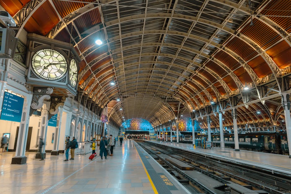 A wide-angle view of the entrance to Paddington Station, featuring a large, curved glass and metal archway with intricate decorative ironwork. The station's name, 'Paddington Station,' is displayed on a white horizontal panel just below the arch. Above the arch, a coat of arms with shields and a crown is visible, mounted at the apex of the structure. The station's roof extends into a covered area with a metal framework and glass panels, allowing natural light to illuminate the interior. The image captures a clear, partly cloudy sky with patches of blue, reflecting in the glass and contrasting with the station's metallic and glass architecture. This setting relates to house removals and moving services offered by Man and Van Paddington, emphasizing the importance of efficient transportation and logistical planning during home relocation, especially when managing loading processes and furniture transport under clear, well-lit conditions.