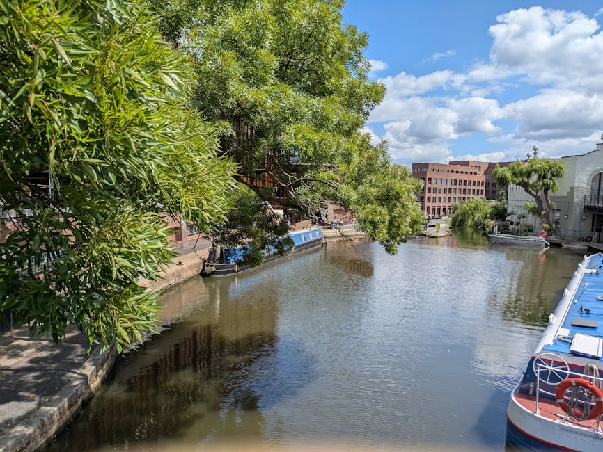 A scenic daytime view of a narrow canal lined with trees and residential buildings, including a white building with balconies and a modern red-brick structure, with a partly cloudy sky overhead. Several boats are moored along the canal's edge, some covered with blue tarpaulins and secured with ropes, while others are open. In the foreground, a cobbled walkway with greenery on the left contrasts with the water. The scene captures a tranquil setting typical of a residential area, suitable for house removals and moving services such as packing, furniture transport, and loading processes. This image is relevant for illustrating home relocation activities near water-based environments, as managed by companies like Man and Van Paddington.