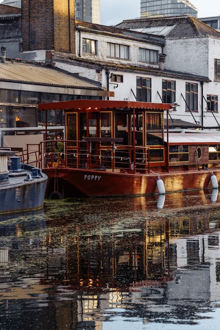 A scenic daytime view of a narrow canal lined with trees and residential buildings, including a white building with balconies and a modern red-brick structure, with a partly cloudy sky overhead. Several boats are moored along the canal's edge, some covered with blue tarpaulins and secured with ropes, while others are open. In the foreground, a cobbled walkway with greenery on the left contrasts with the water. The scene captures a tranquil setting typical of a residential area, suitable for house removals and moving services such as packing, furniture transport, and loading processes. This image is relevant for illustrating home relocation activities near water-based environments, as managed by companies like Man and Van Paddington.