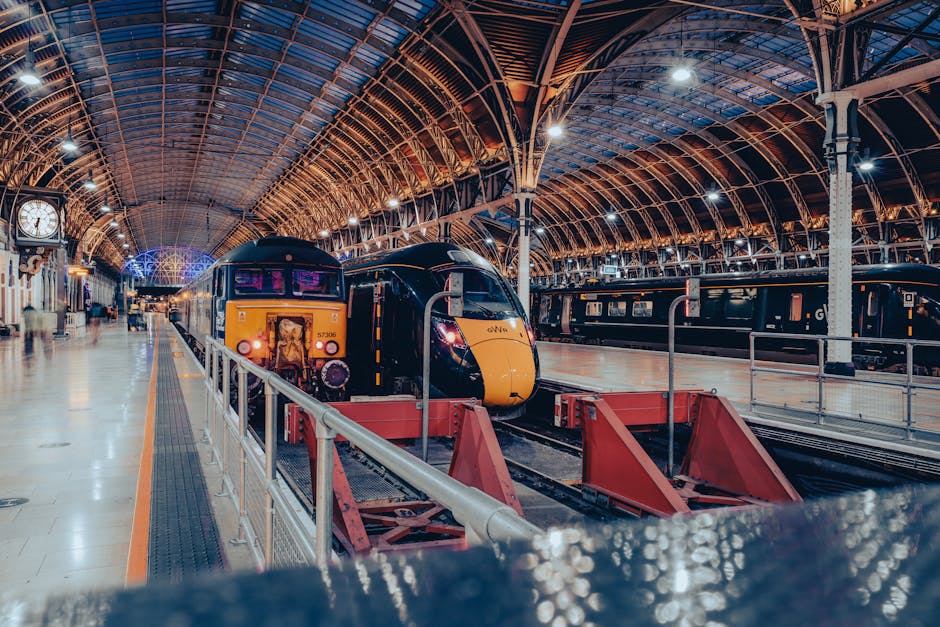 Inside a spacious train station with a high, arched, steel and glass ceiling illuminated by bright overhead lighting. On the platform, a modern yellow and black passenger train is stationary, with visible front headlights and a driver’s cab. To the left, there is a vintage-style clock mounted on the wall, showing the time. The platform is clean, with a few people visible in the background, some walking and others waiting. There are multiple sets of railway tracks, with one track engaged with the train, and others empty. Adjacent to the train, several open boxes and luggage are placed on the platform, possibly prepared for a home relocation or packing process. Man and Van Paddington is providing the service, with a focus on safe and organized furniture transport and packing during house moves, ensuring minimal disruption. The environment is well-lit and orderly, emphasizing efficient moving logistics within a railway station setting, suitable for complex house removals and transport logistics.