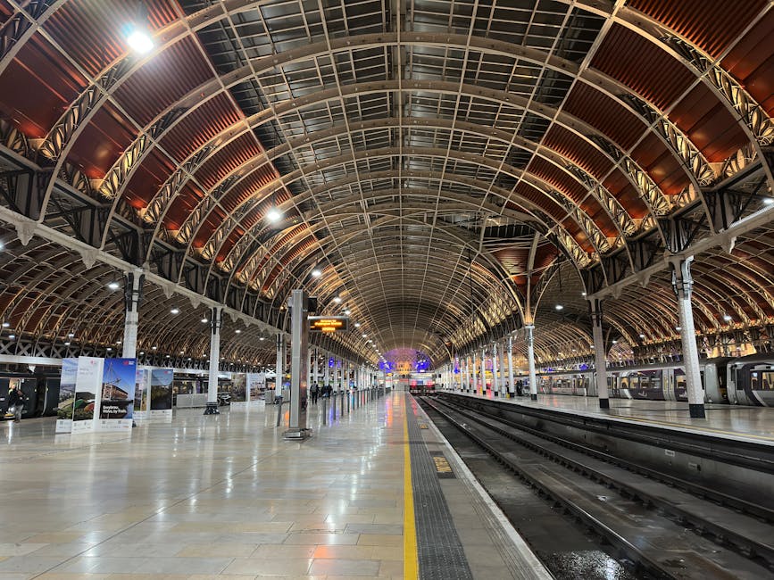 An interior view of a spacious train station with a high, arched ceiling made of intricate metal framework and warm-toned panels. The platform is wide and mostly empty, with polished stone flooring reflecting the ambient lighting. On the right side, several modern trains with dark windows are stationary alongside the platform. To the left, there are boarded-up advertising displays and signs, some showing images and information. A few passengers are visible in the distance, walking or waiting. Overhead, digital departure boards display train times and destinations in bright text. The station's lighting is bright yet diffused, highlighting the architectural details of the vaulted ceiling. The scene captures the typical environment for home relocation or furniture transport, where the clear platform space and train access are essential for moving large items, often requiring loading equipment like trolleys or straps. Man and Van Paddington operates in such environments to assist with packing, loading, and transport during house removals within Paddington W2, ensuring compliance with parking and access rules for vans.