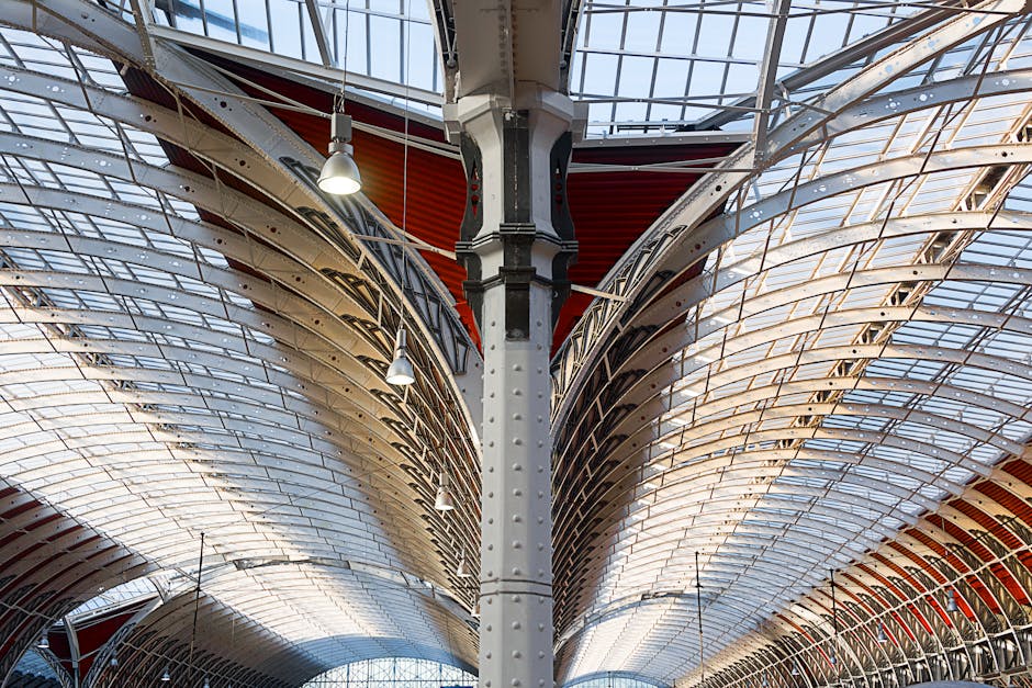 A view of the intricately designed ceiling of a modern transport hub or station, featuring large, curved metal and glass panels that create an expansive and airy environment. The ceiling structure includes numerous metal beams and trusses painted in white and red, supporting the translucent glass roof that allows natural light to fill the space. Several overhead lamps hang from the ceiling, providing additional illumination. The image emphasizes the architectural details and the spacious interior, possibly an area used for passenger movement or freight loading. Man and Van Paddington may utilize such spaces for loading and unloading furniture or moving boxes during home relocation processes, with the environment suitable for the logistics of packing and furniture transport, illustrating the importance of well-structured facilities in efficient removals services.