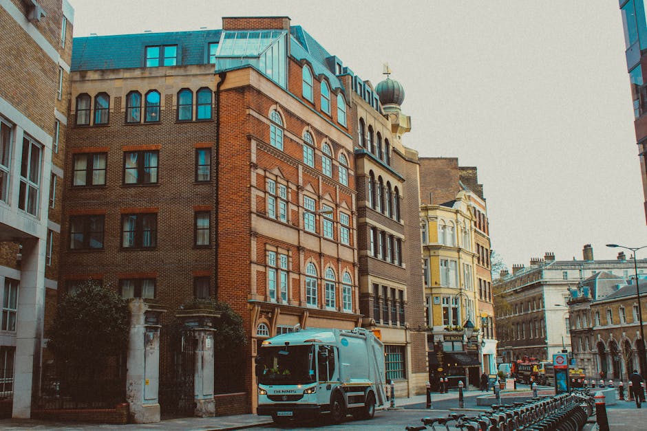 A white street cleaning van parked on the pavement in front of historic and modern multi-storey buildings in Paddington, London. The buildings are constructed with red and yellow brick, featuring large arched windows, decorative cornices, and a small dome on the roof with a cross on top. The street scene includes bollards, bicycles, street lamps, and a bus stop, with a clear sky overhead. The van, associated with furniture transport and home relocation services, is situated near an entrance to a property, possibly during a house removal or packing process, with nearby cardboard boxes and moving equipment visible on the sidewalk, reflecting the ongoing logistics of a house move coordinated by Man and Van Paddington.