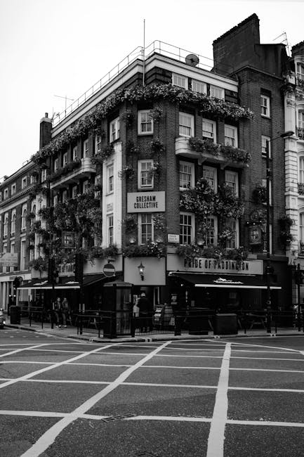 Black and white photograph of a multi-storey building on a city street corner, featuring several balconies adorned with hanging flower baskets. The building has a ground-floor pub or restaurant with outdoor seating, visible tables and chairs, and a sign reading 'Pride of Paddington.' In front of the building, there is a pedestrian crossing with marked white lines on the road. The street appears to be busy, with street lamps and signage visible. The image illustrates a typical urban environment suitable for home removal or relocation services, with no visible furniture or moving equipment present.